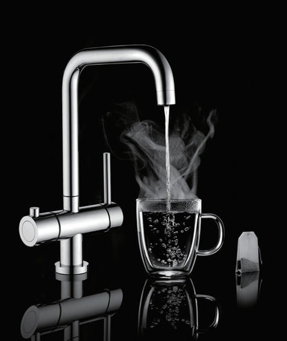 A modern chrome double-handle faucet with boiling water running into a clear glass mug, shown on a black reflective background with a tea bag beside the mug.