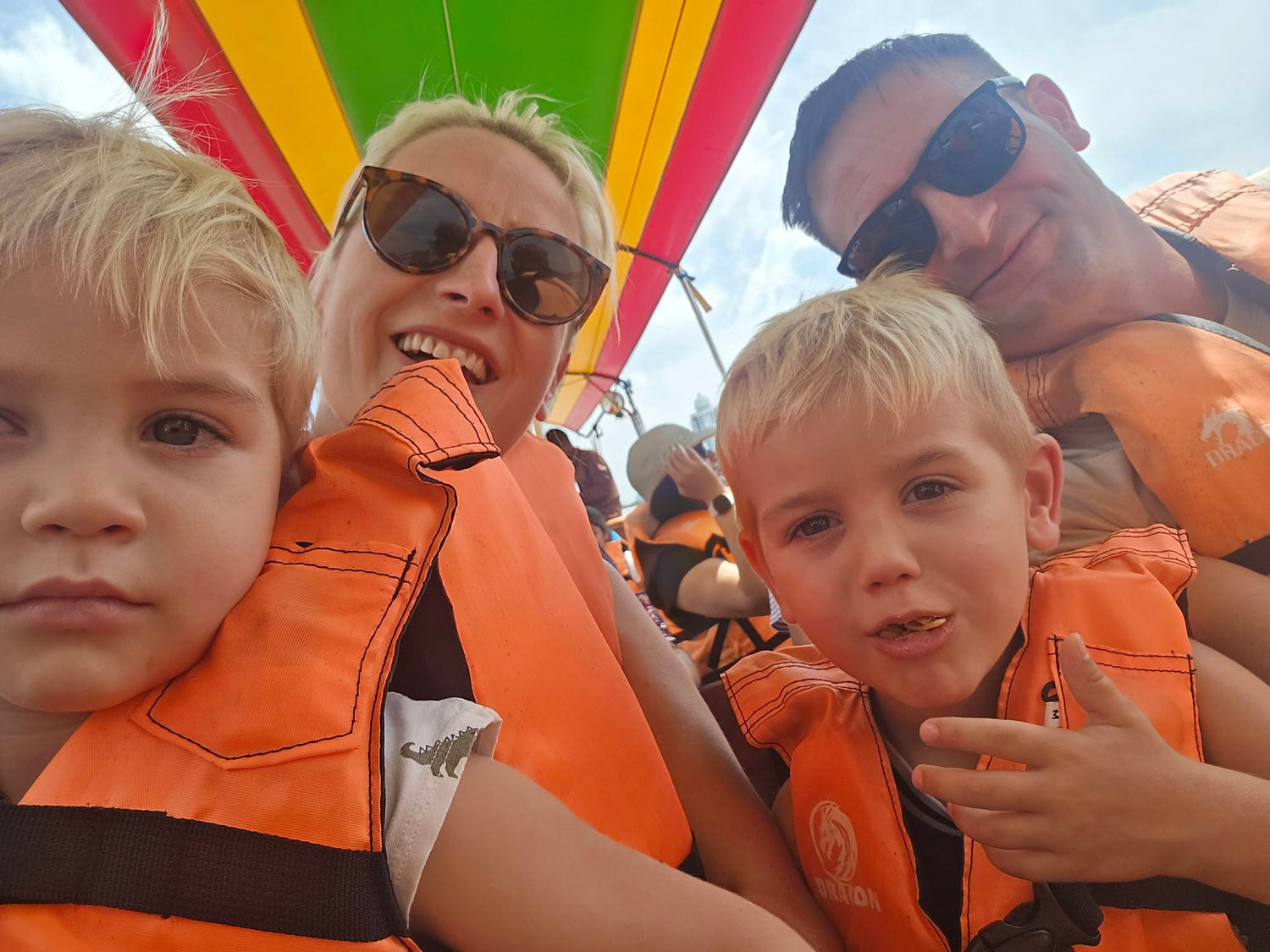 Family with two children wearing orange life vests on a boat, with colorful striped canopy in the background.