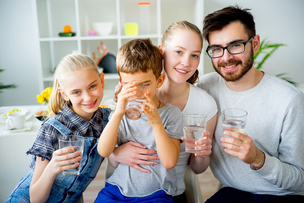 A family of four sitting together indoors, each holding a clear glass of water. The father wears glasses and a light gray long-sleeve shirt, the mother wears a white top, the daughter wears denim overalls over a plaid shirt, and the son wears a gray t-shirt. All drinking chilled filtered water.